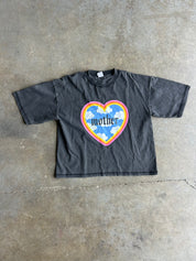 A faded black short-sleeve t-shirt laid flat on a concrete floor, featuring a colorful heart graphic on the chest with the word 'mother' inside