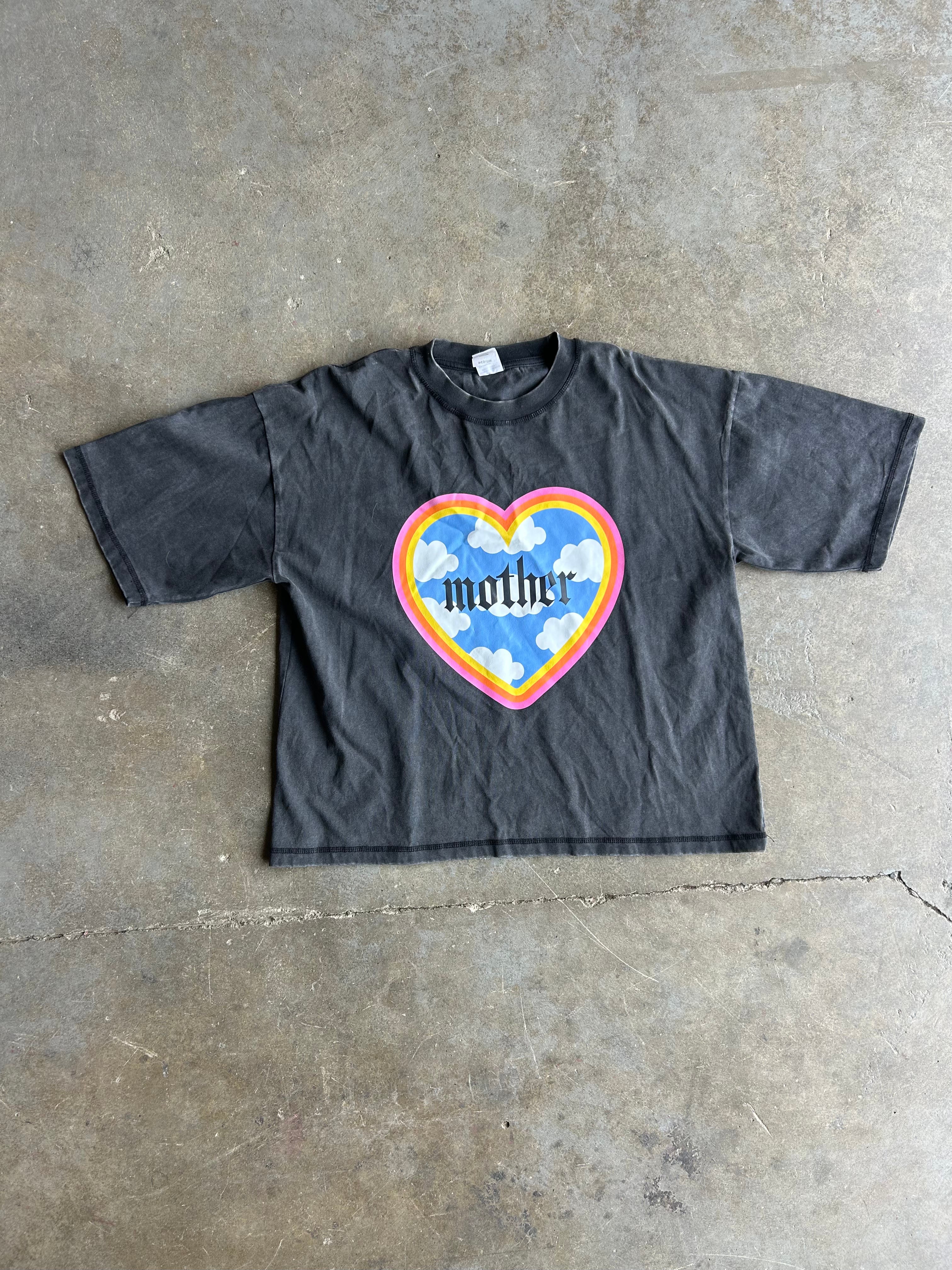 A faded black short-sleeve t-shirt laid flat on a concrete floor, featuring a colorful heart graphic on the chest with the word 'mother' inside