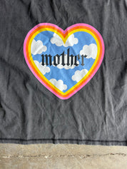Close up of a faded black short-sleeve t-shirt laid flat on a concrete floor, featuring a colorful heart graphic on the chest with the word 'mother' inside
