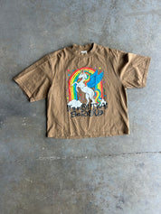 A brown short-sleeve t-shirt laid flat on a gray concrete floor, featuring a colorful graphic of a white unicorn with a blue wing under a rainbow, with the text 'It's Not That Serious' printed below