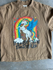 Close up of a brown short-sleeve graphic t-shirt featuring a white winged unicorn in front of a rainbow, with the text 'It's not that serious' printed below. The shirt is laid flat on a gray concrete floor