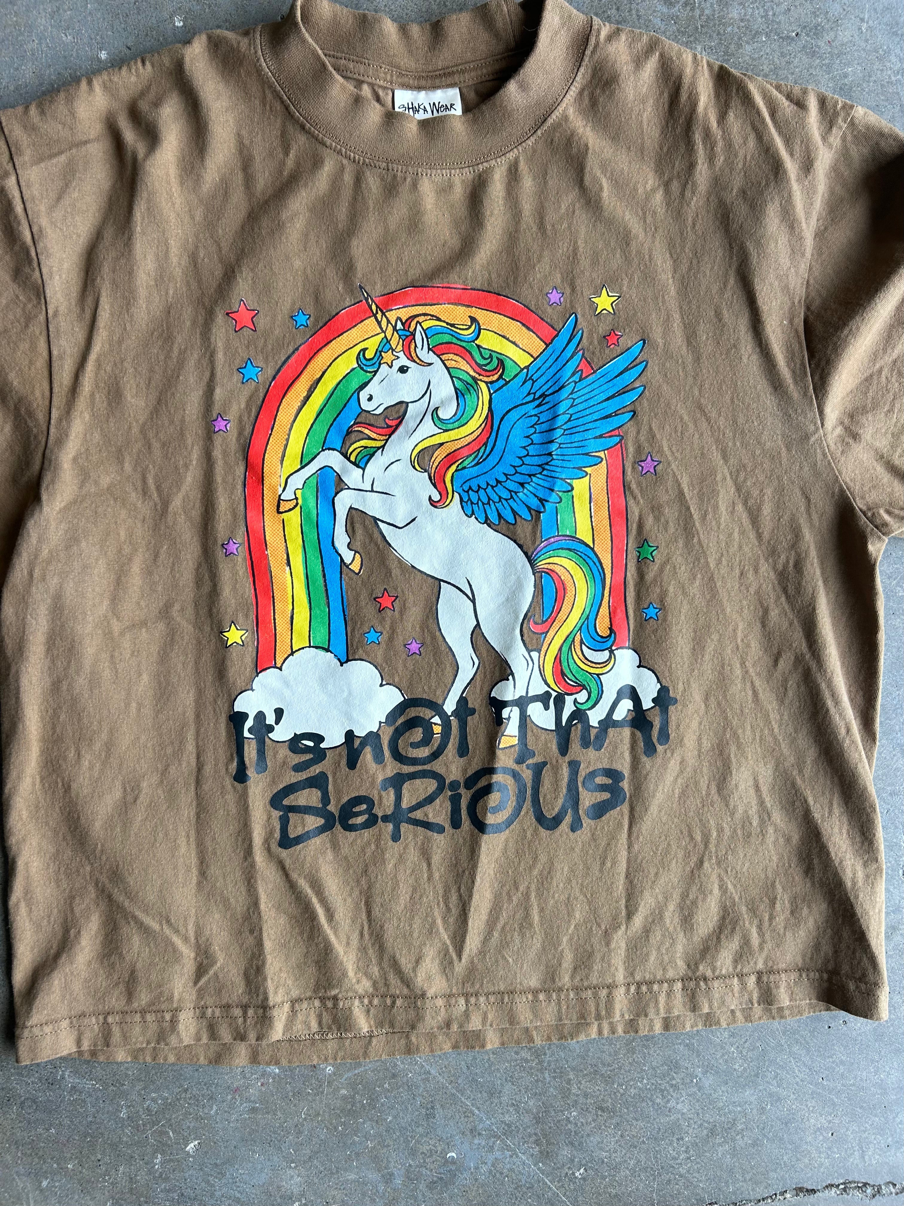 Close up of a brown short-sleeve graphic t-shirt featuring a white winged unicorn in front of a rainbow, with the text 'It's not that serious' printed below. The shirt is laid flat on a gray concrete floor