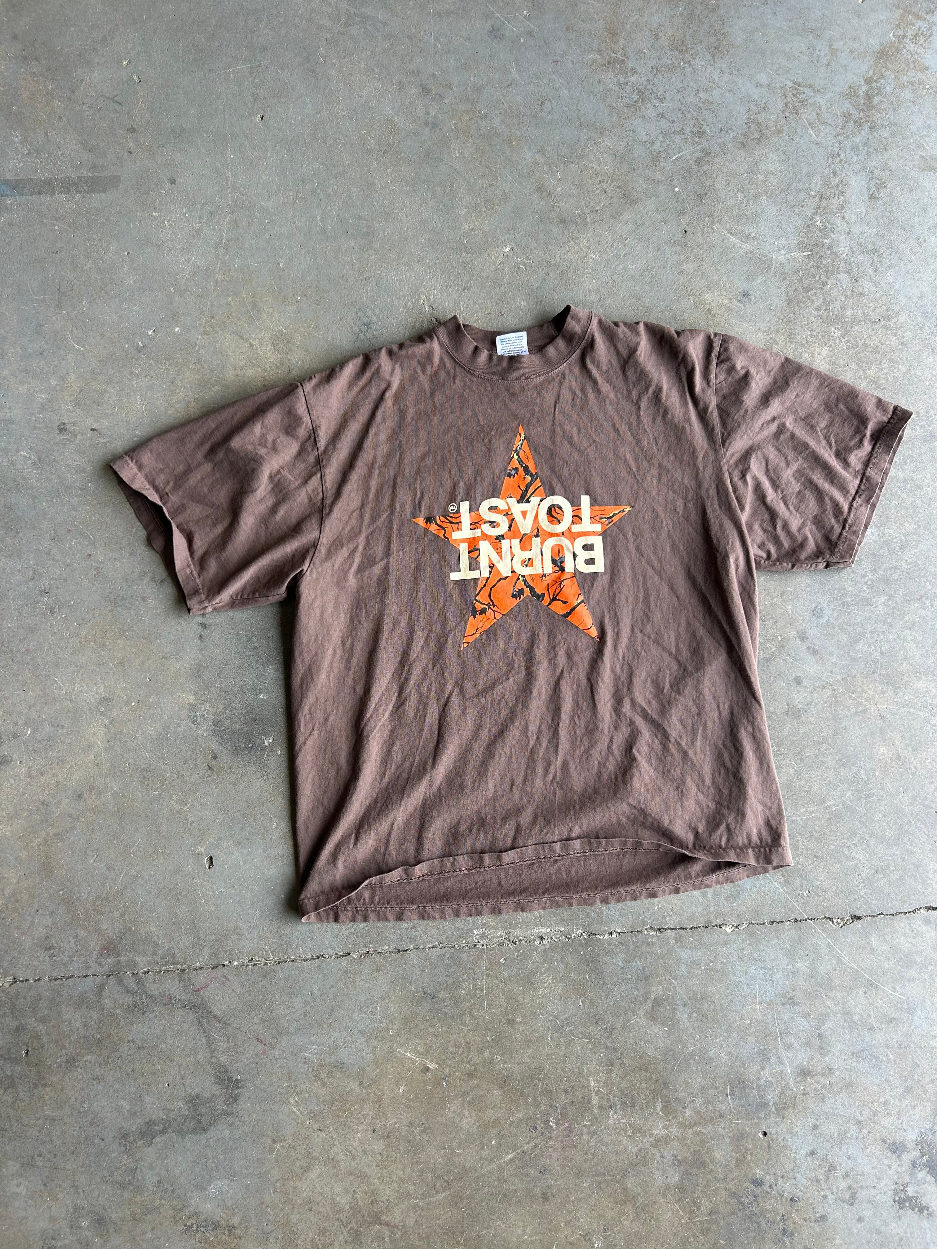 A brown short-sleeve t-shirt laid flat on a concrete floor, featuring an orange star graphic with the words 'BURN TOAST' in white text