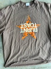 A faded brown short-sleeve t-shirt laid flat on a gray concrete surface, featuring a large orange star graphic with a camouflage pattern and the words 'BURNT TOAST' printed in white across the center