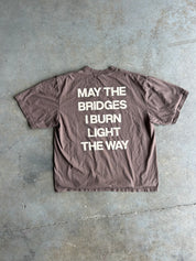 Back of a brown tee shirt with the text 'MAY THE BRIDGES I BURN LIGHT THE WAY' in cream on a concrete surface