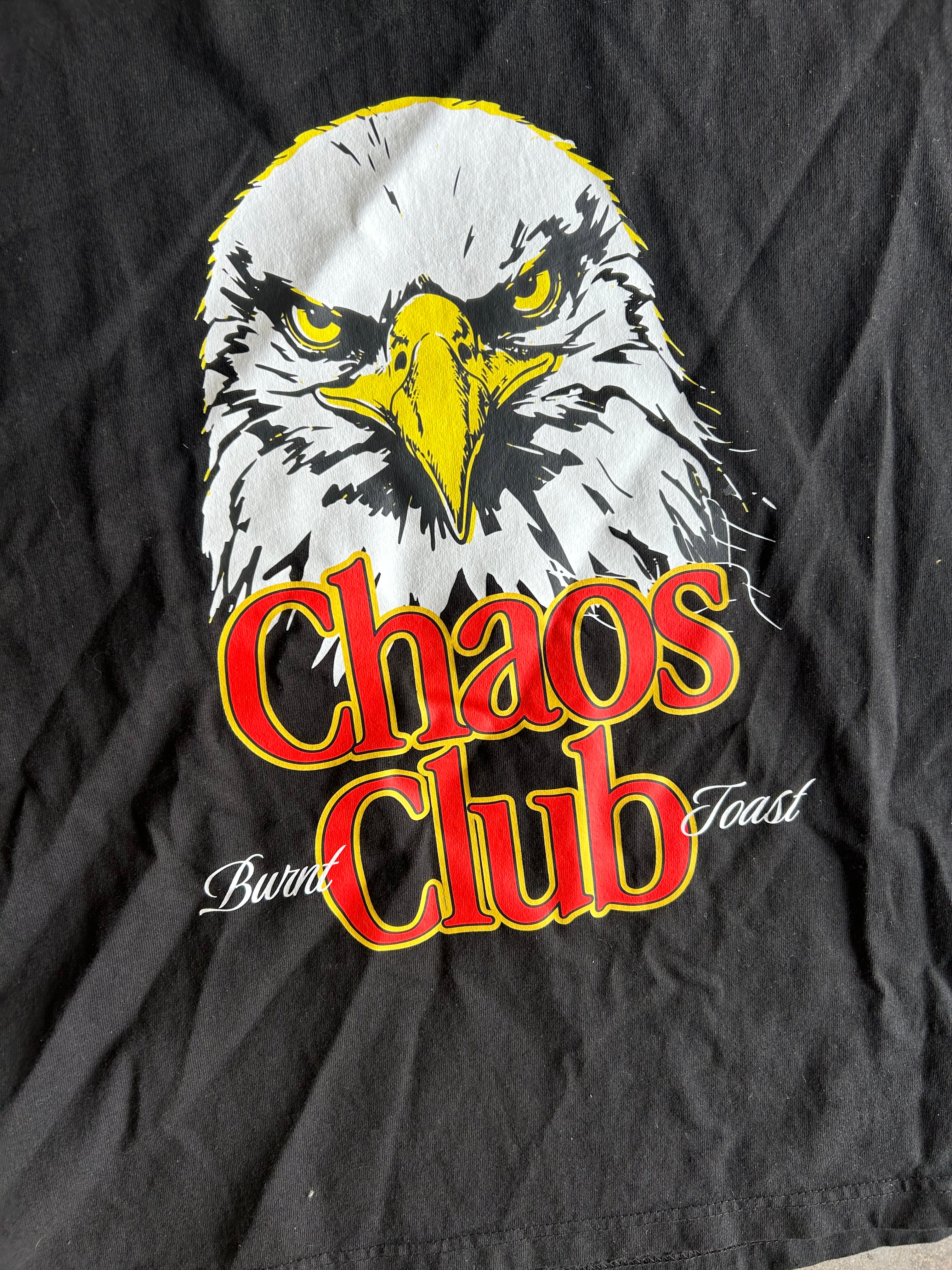 A close-up of a black graphic t-shirt featuring a bald eagle illustration and the words 'Chaos Club' in red with yellow outlines, along with 'Burnt Toast' in white script.