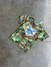 A camouflage short-sleeve t-shirt with a colorful heart graphic on the chest, laid flat on a concrete floor. The graphic features a blue heart with the word 'mother' in black script, surrounded by a pink, orange, and yellow border.