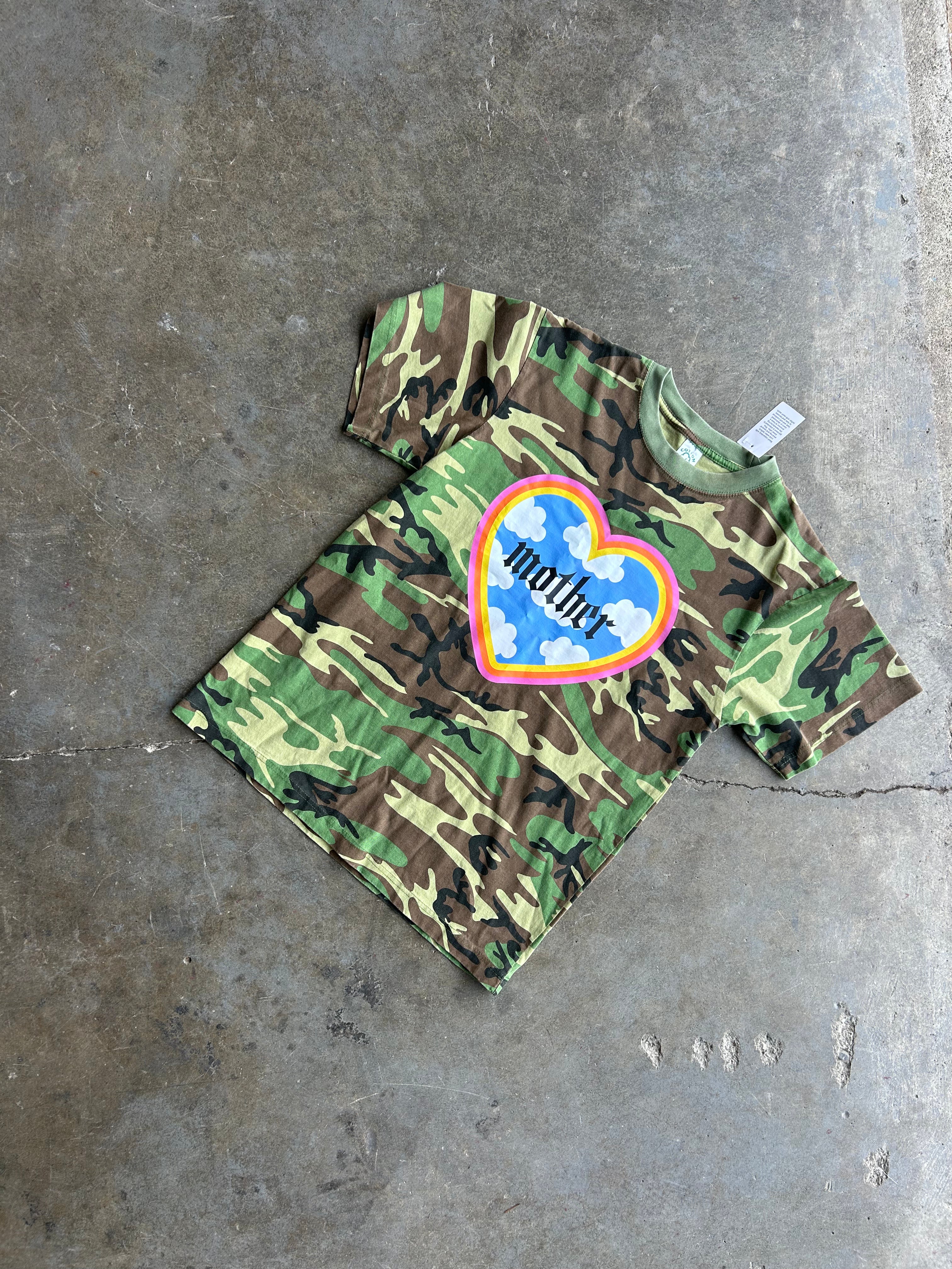 A camouflage short-sleeve t-shirt with a colorful heart graphic on the chest, laid flat on a concrete floor. The graphic features a blue heart with the word 'mother' in black script, surrounded by a pink, orange, and yellow border.