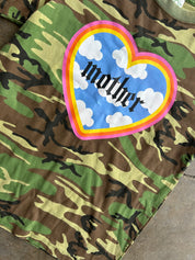 A close-up of a camouflage t-shirt featuring a large heart-shaped graphic on the chest. The graphic has a blue sky and white clouds background with the word 'mother' written in black gothic font, surrounded by a pink, orange, and yellow border.