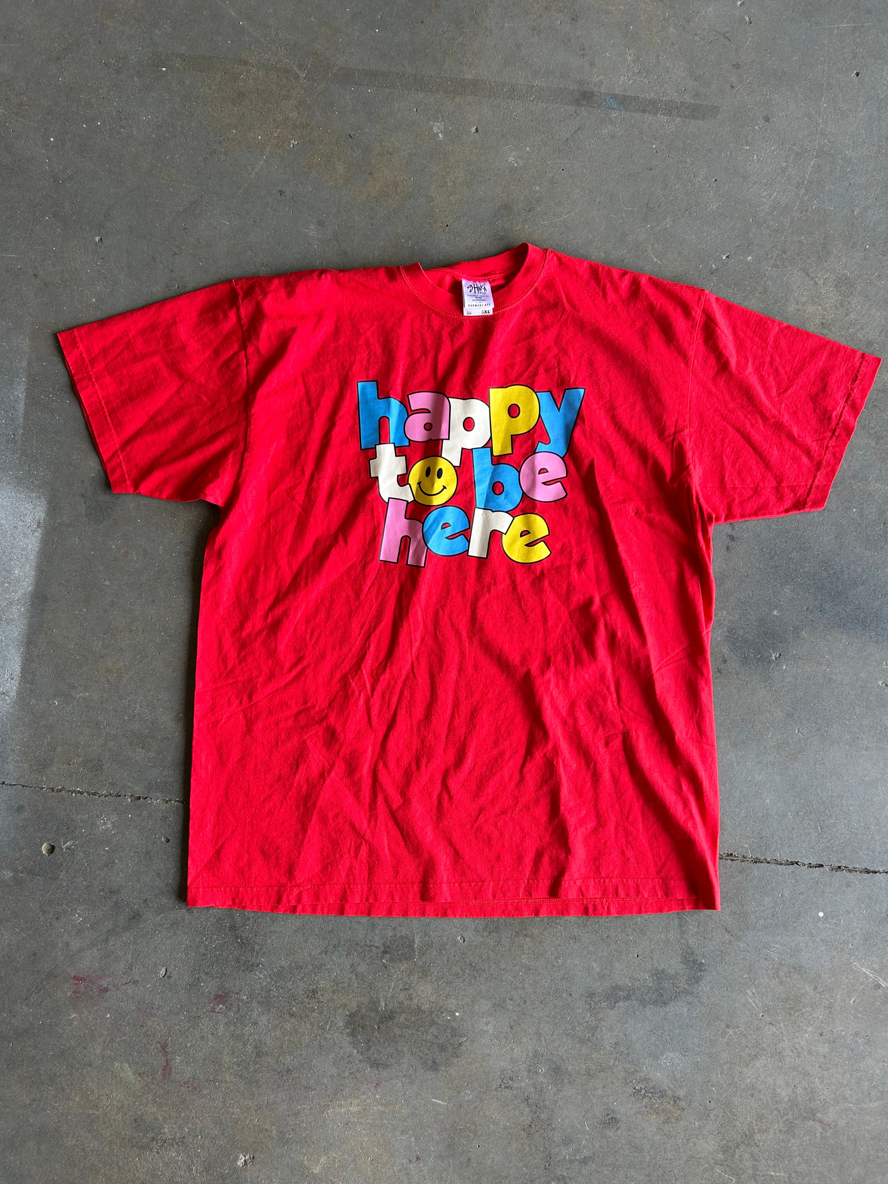 Red graphic tee with Happy to Be Here text on a concrete surface