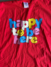 A red short-sleeve t-shirt featuring a colorful graphic that reads 'happy to be here' in bubble letters with a smiley face, laid flat.