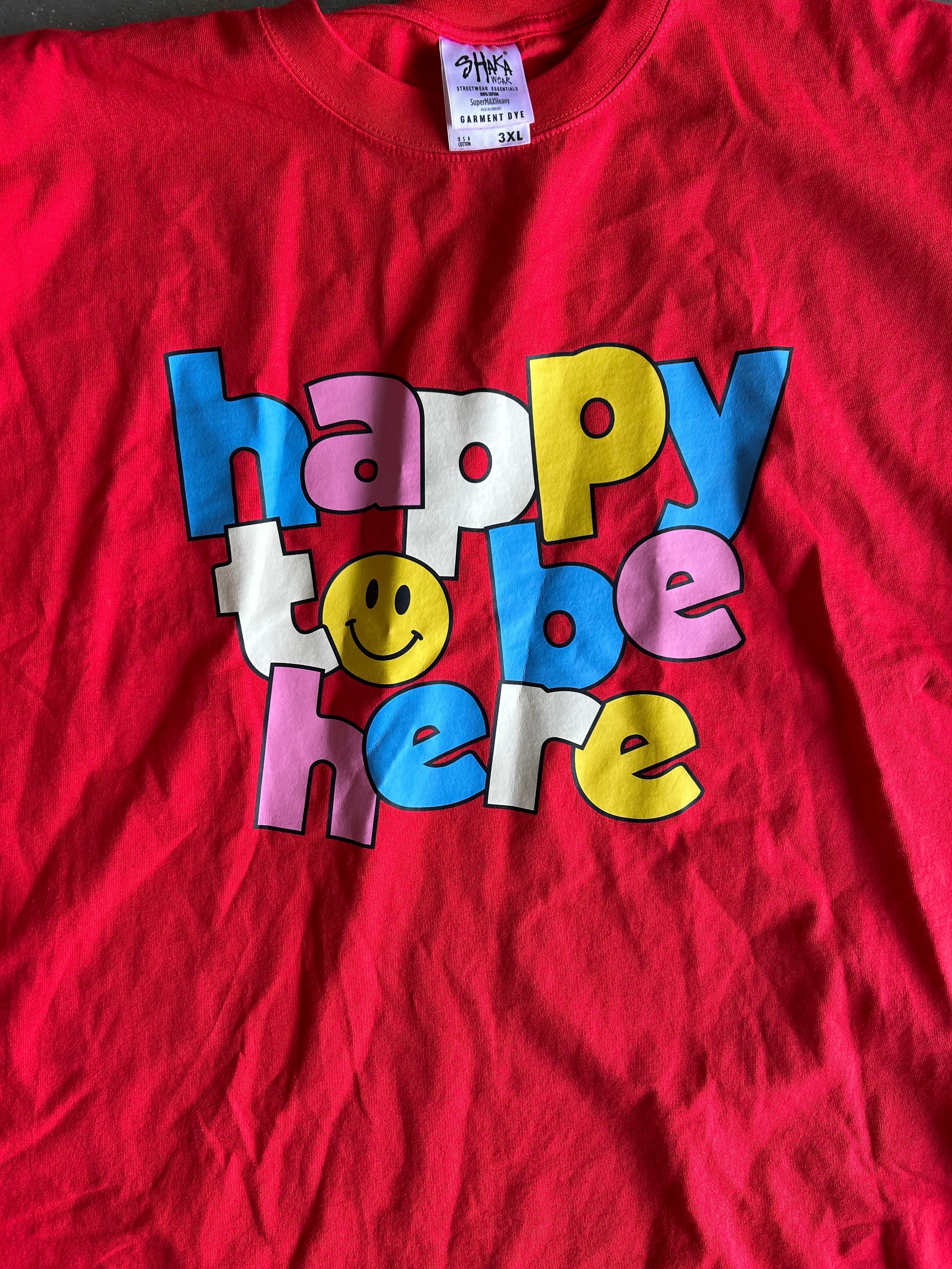 A red short-sleeve t-shirt featuring a colorful graphic that reads 'happy to be here' in bubble letters with a smiley face, laid flat.