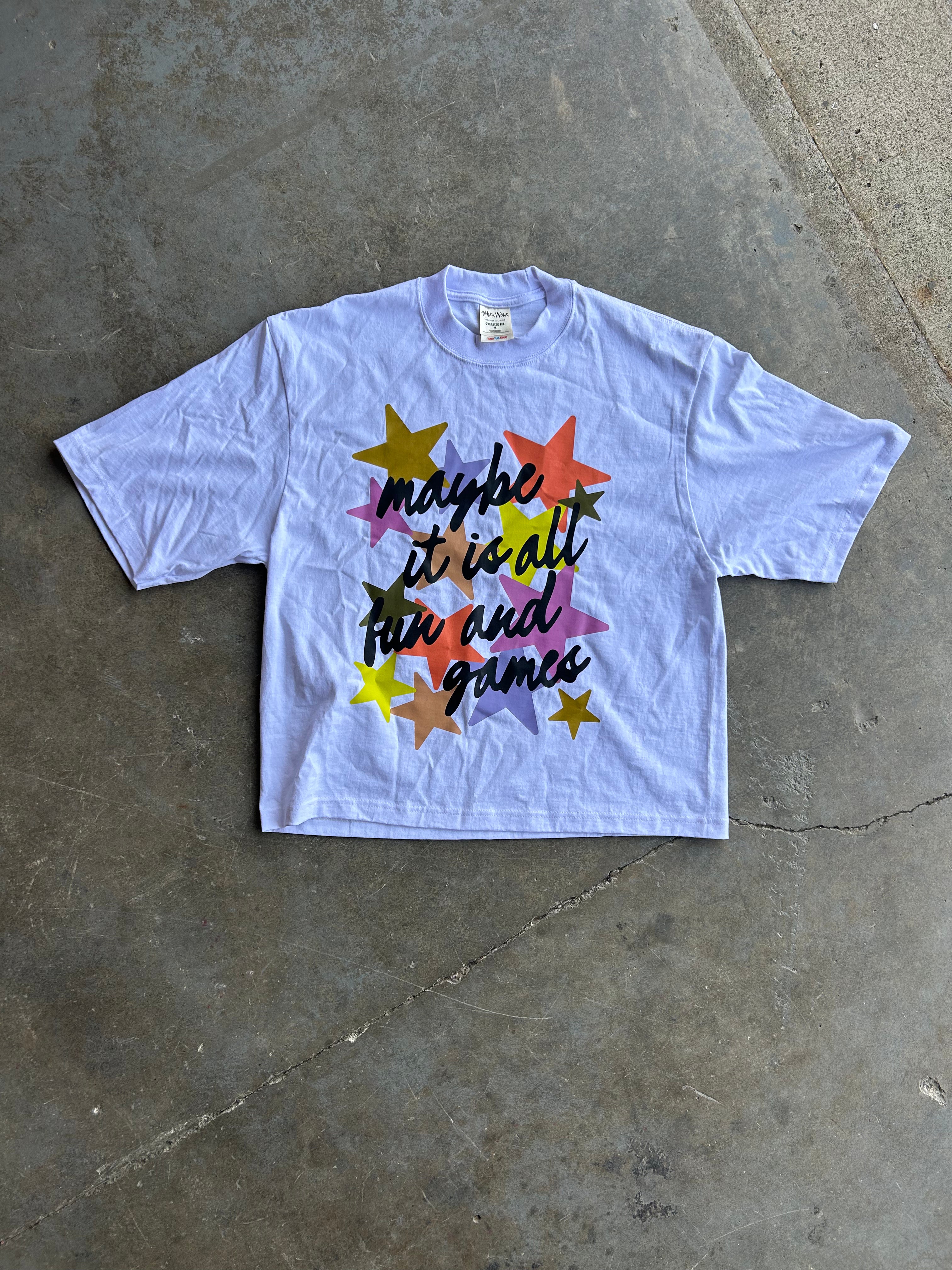 A light blue short-sleeve graphic t-shirt laid flat on a gray concrete floor. The shirt features a colorful graphic with yellow, orange, pink, purple, and blue stars surrounding the black cursive text 'maybe it is all fun and games'.