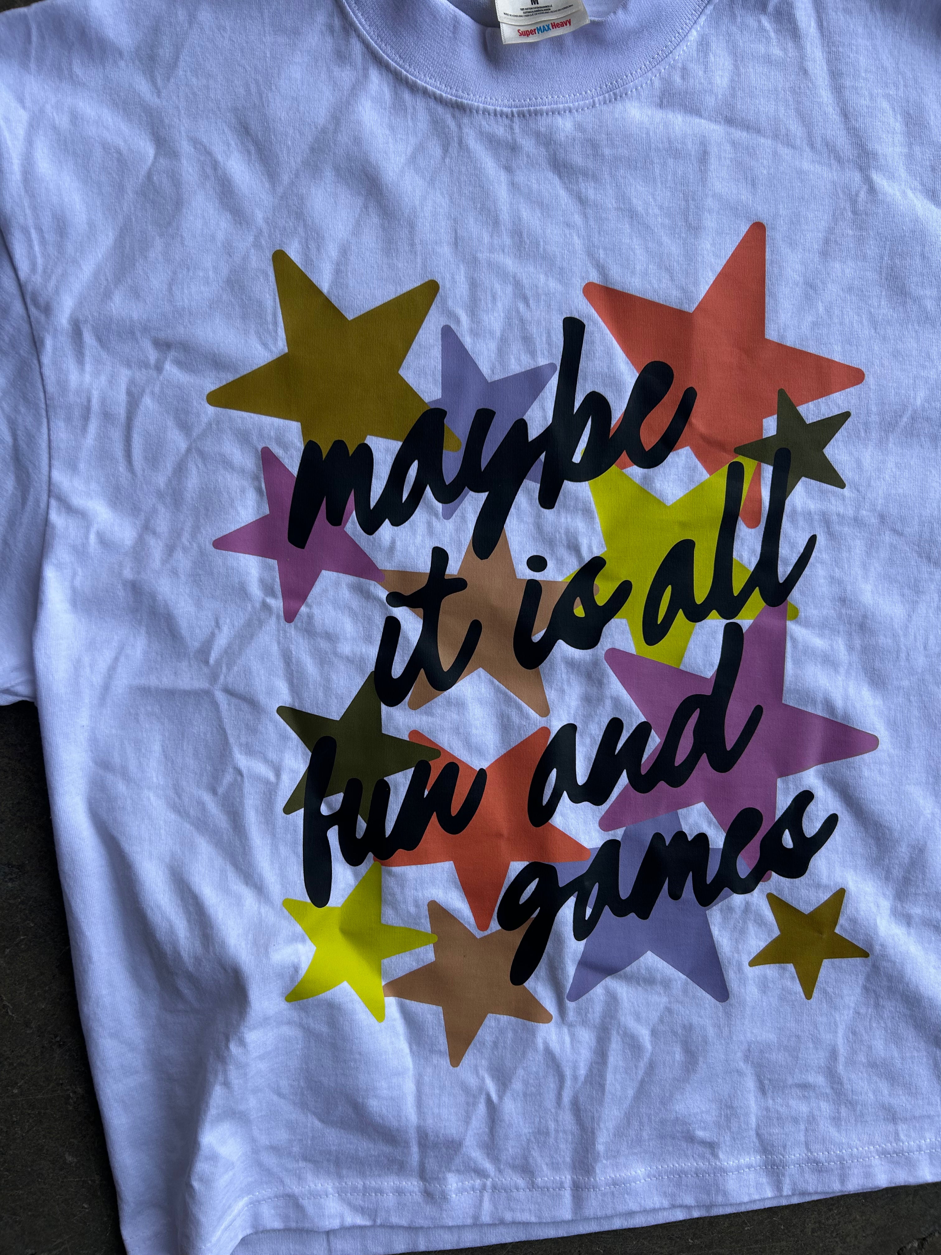 A light blue crewneck t-shirt featuring a colorful graphic of overlapping stars in yellow, orange, purple, brown, and green, with the black cursive text 'maybe it is all fun and games' printed across the front.