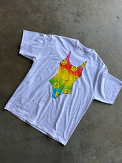 A white short-sleeve t-shirt laid flat on a gray concrete floor, featuring a colorful rainbow gradient graphic in the center with yellow smiley faces.