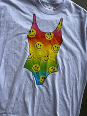 A light gray crewneck t-shirt featuring a large graphic print of a rainbow-striped bodysuit patterned with yellow smiley faces, laid flat on a dark surface.