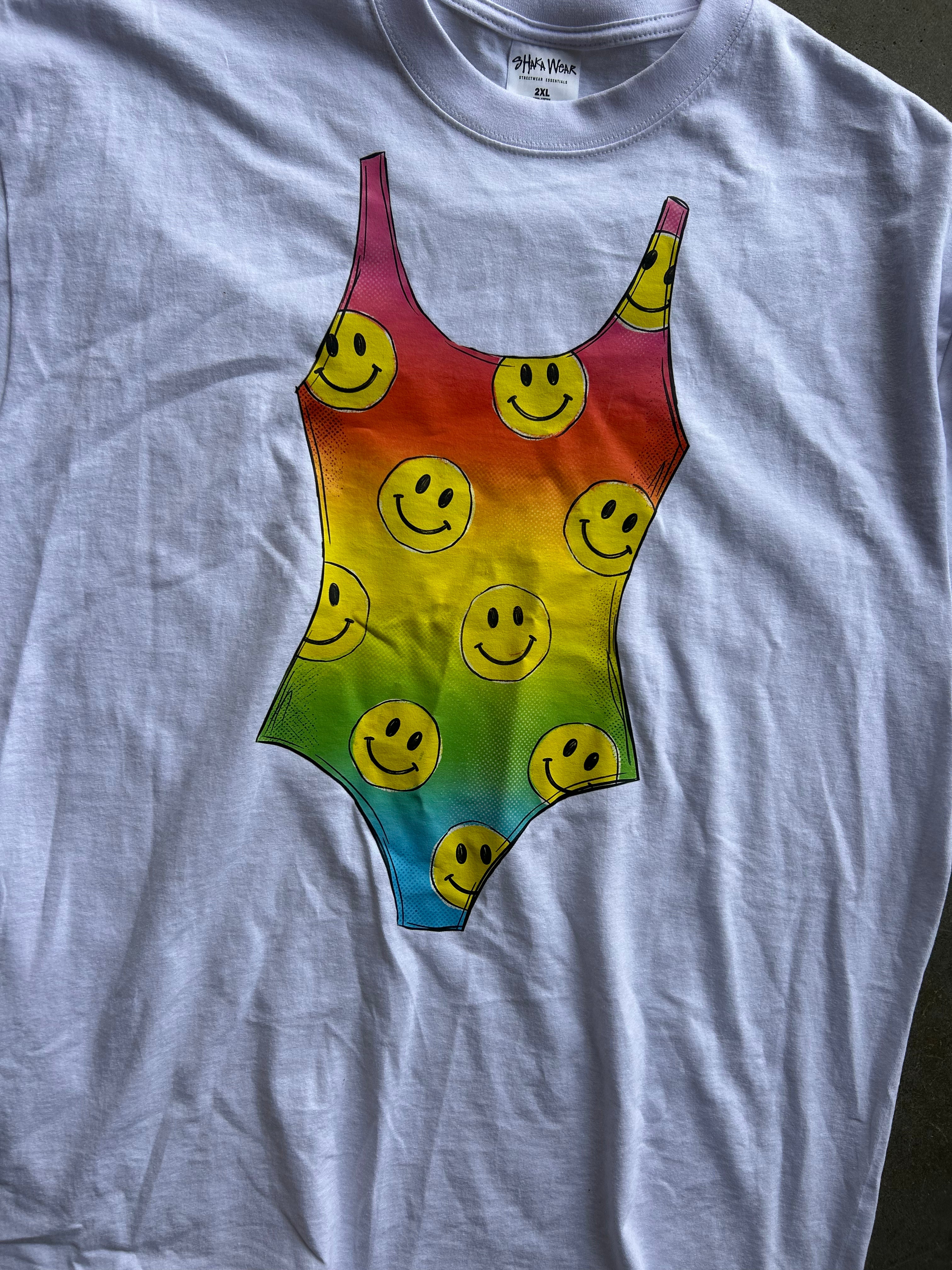 A light gray crewneck t-shirt featuring a large graphic print of a rainbow-striped bodysuit patterned with yellow smiley faces, laid flat on a dark surface.