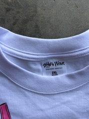 Close-up of the inside collar of a white crewneck t-shirt, showing the 'SHAKA WEAR' brand label and '2XL' size text, set against a textured gray concrete background.