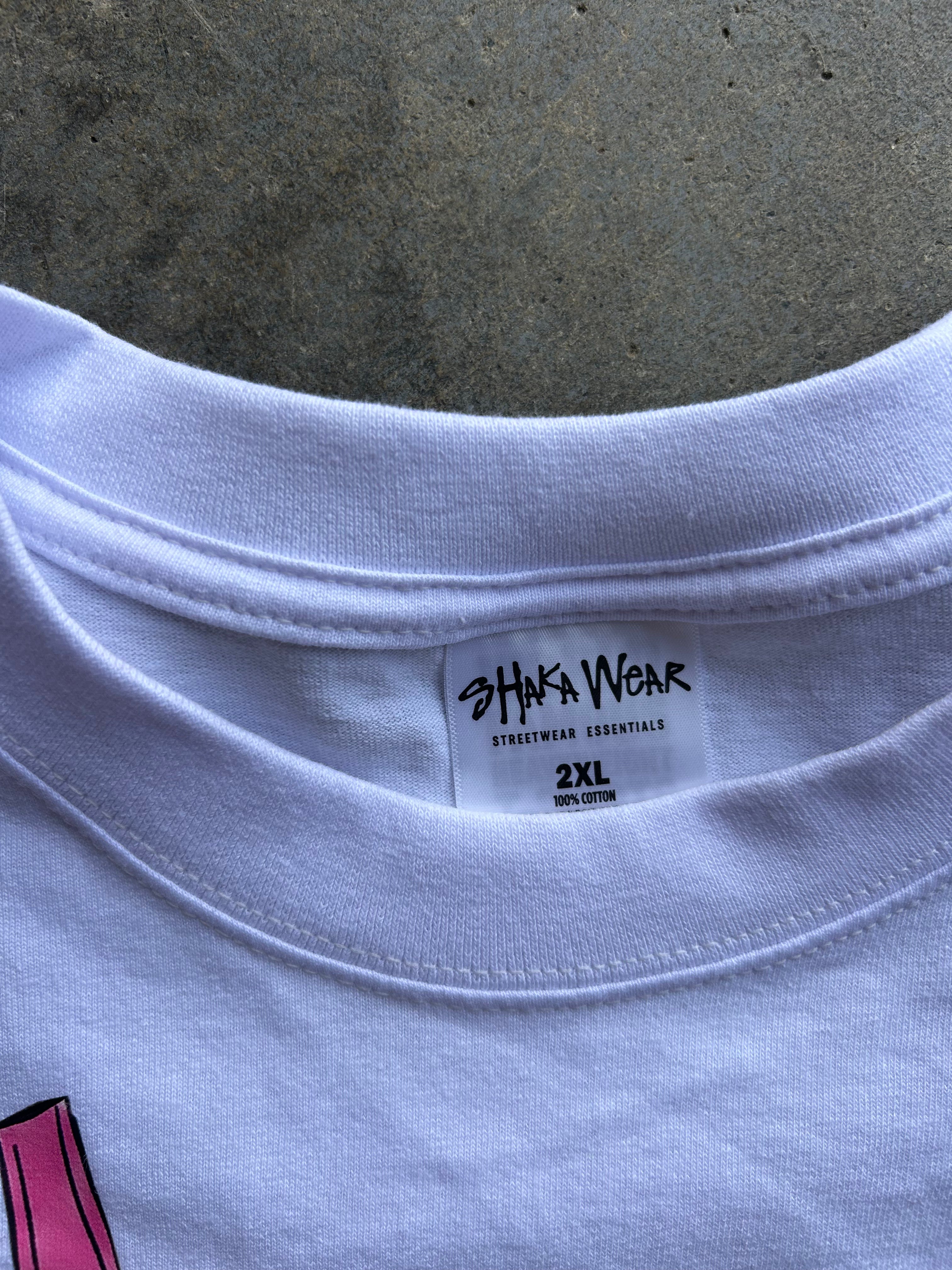 Close-up of the inside collar of a white crewneck t-shirt, showing the 'SHAKA WEAR' brand label and '2XL' size text, set against a textured gray concrete background.