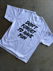 A white short-sleeve t-shirt with the phrase 'DON'T FORGET TO HAVE FUN' printed in black stylized text on the back, laid flat on a gray concrete floor.