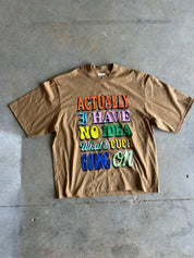 A brown short-sleeve graphic t-shirt laid flat on a gray concrete floor. The shirt features a colorful retro-style text graphic that reads 'ACTUALLY I HAVE NO IDEA What's even GOING ON' in various vibrant colors.