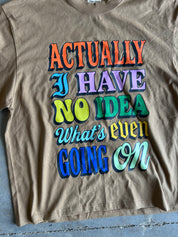 A tan crewneck t-shirt featuring a colorful retro-style graphic text that reads 'ACTUALLY I HAVE NO IDEA What's even GOING ON' in orange, blue, purple, green, yellow, and light blue lettering with dark drop shadows, laid flat on a gray concrete floor.