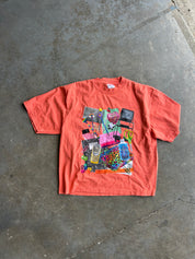 A coral orange short-sleeve t-shirt with a colorful graphic collage print on the front, laid flat on a gray concrete floor.