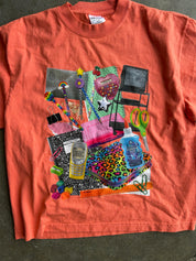 A coral orange short-sleeve t-shirt laid flat on a dark gray concrete floor, featuring a large, colorful collage graphic of school supplies and everyday objects on the front.