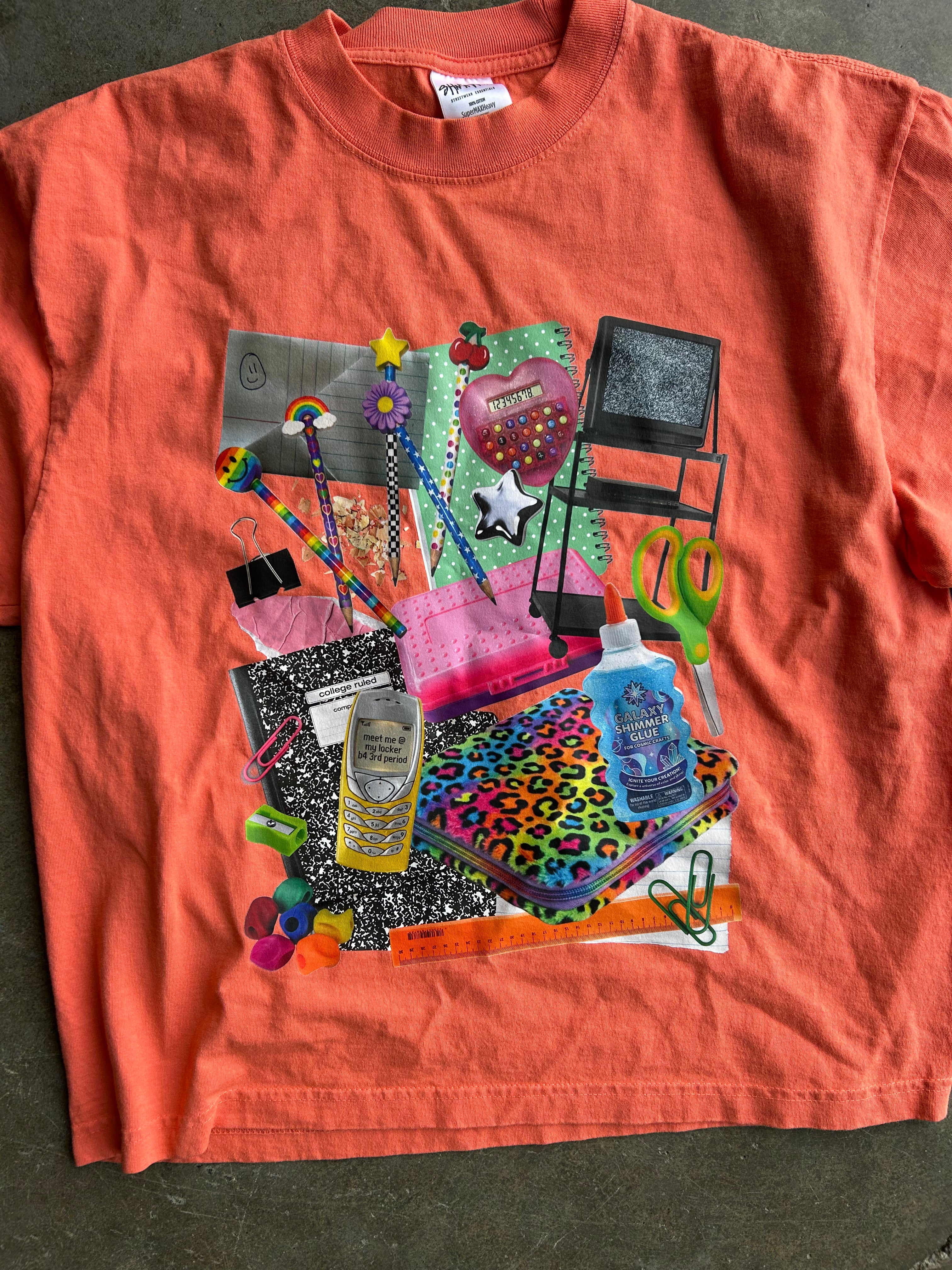 A coral orange short-sleeve t-shirt laid flat on a dark gray concrete floor, featuring a large, colorful collage graphic of school supplies and everyday objects on the front.