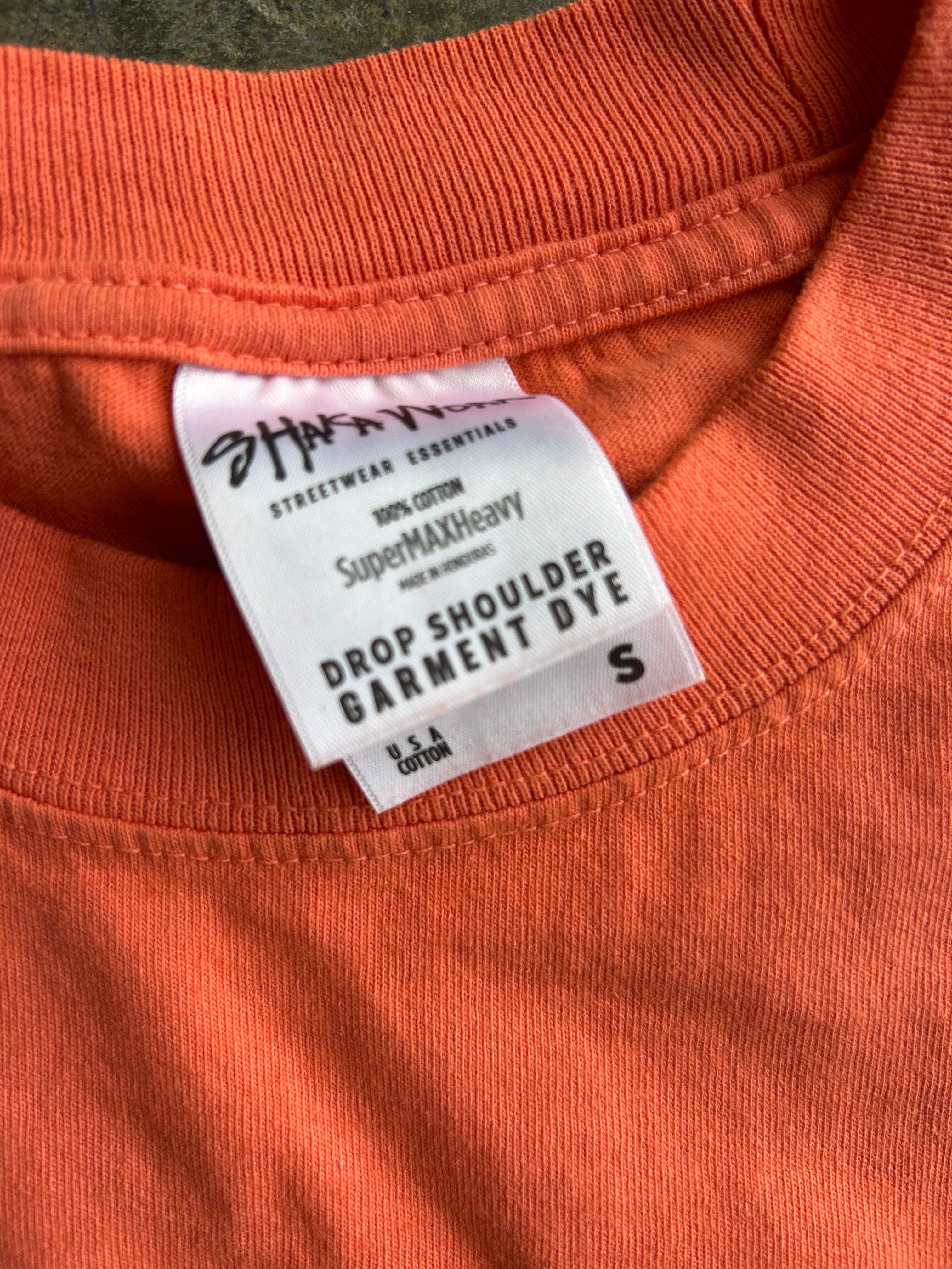 Close-up of the inside collar of an orange crewneck garment, showing a white tag that reads 'Streetwear Essentials SuperMAX Heavy Drop Shoulder Garment Dye' and size 'S'.