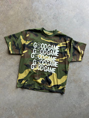 A short-sleeve crewneck t-shirt with a green, brown, and yellow camouflage pattern, featuring the text 'GOOD GAME' repeated five times in white with smiley faces integrated into the letters. The shirt is laid flat on a textured gray concrete floor.