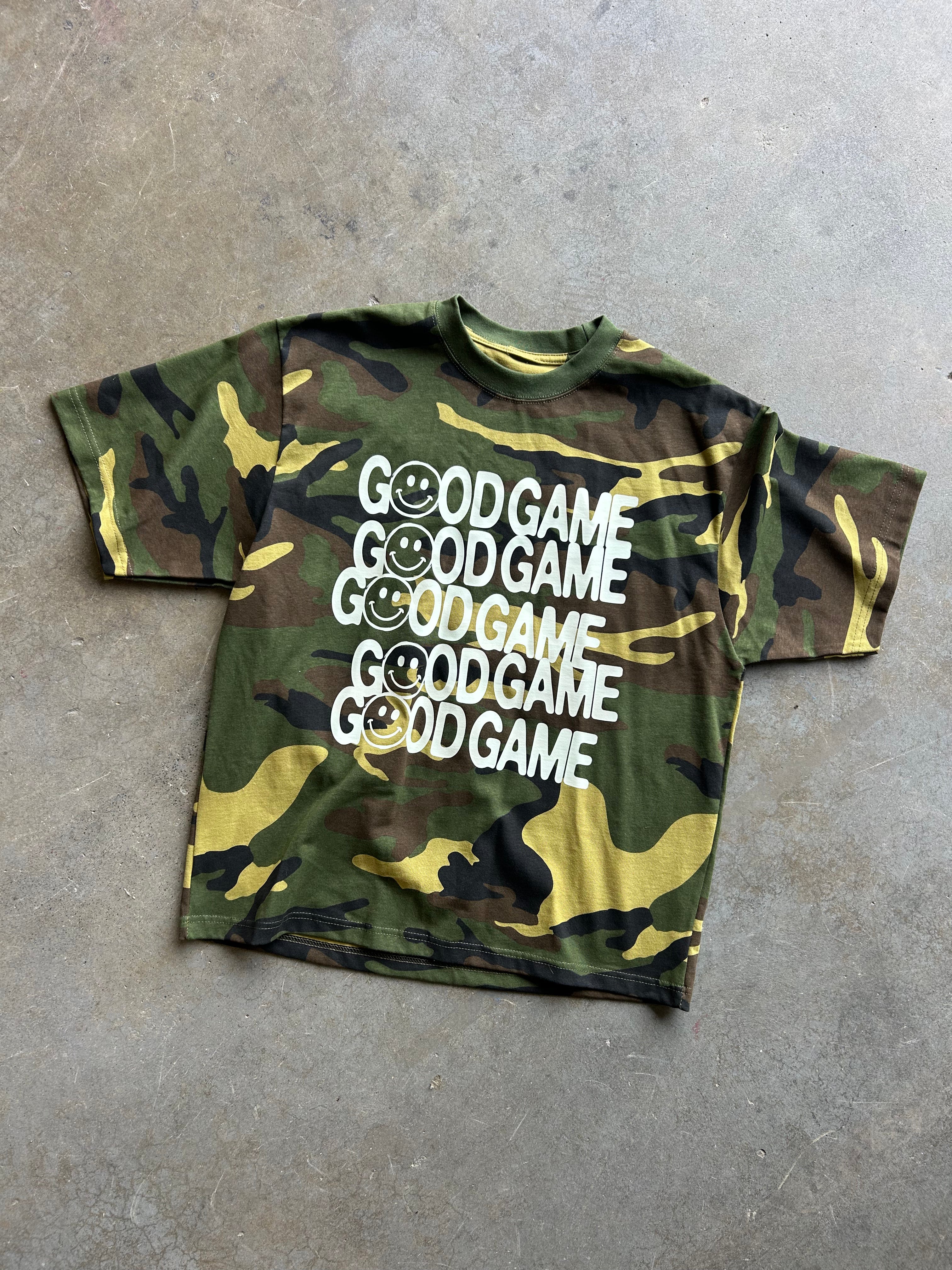 A short-sleeve crewneck t-shirt with a green, brown, and yellow camouflage pattern, featuring the text 'GOOD GAME' repeated five times in white with smiley faces integrated into the letters. The shirt is laid flat on a textured gray concrete floor.