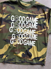 A camouflage t-shirt with a green, brown, and tan camo pattern, featuring the text 'GOOD GAME' repeated vertically in white, with a smiley face graphic inside the letter 'O'.