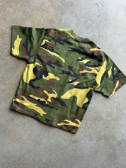 A short-sleeve camouflage t-shirt laid flat on a textured gray concrete floor. The shirt features a classic woodland camo pattern with green, brown, black, and yellow shapes.