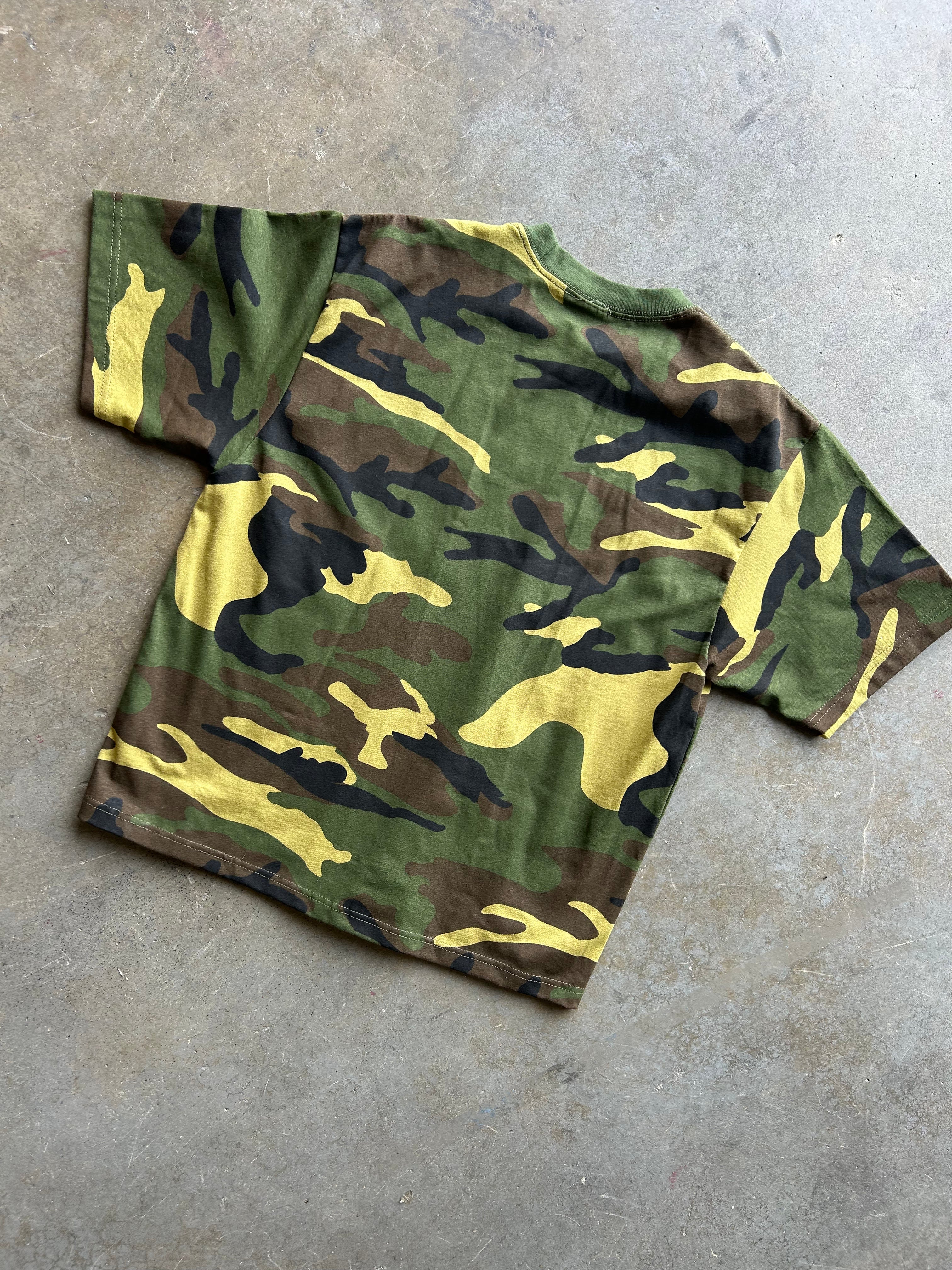 A short-sleeve camouflage t-shirt laid flat on a textured gray concrete floor. The shirt features a classic woodland camo pattern with green, brown, black, and yellow shapes.