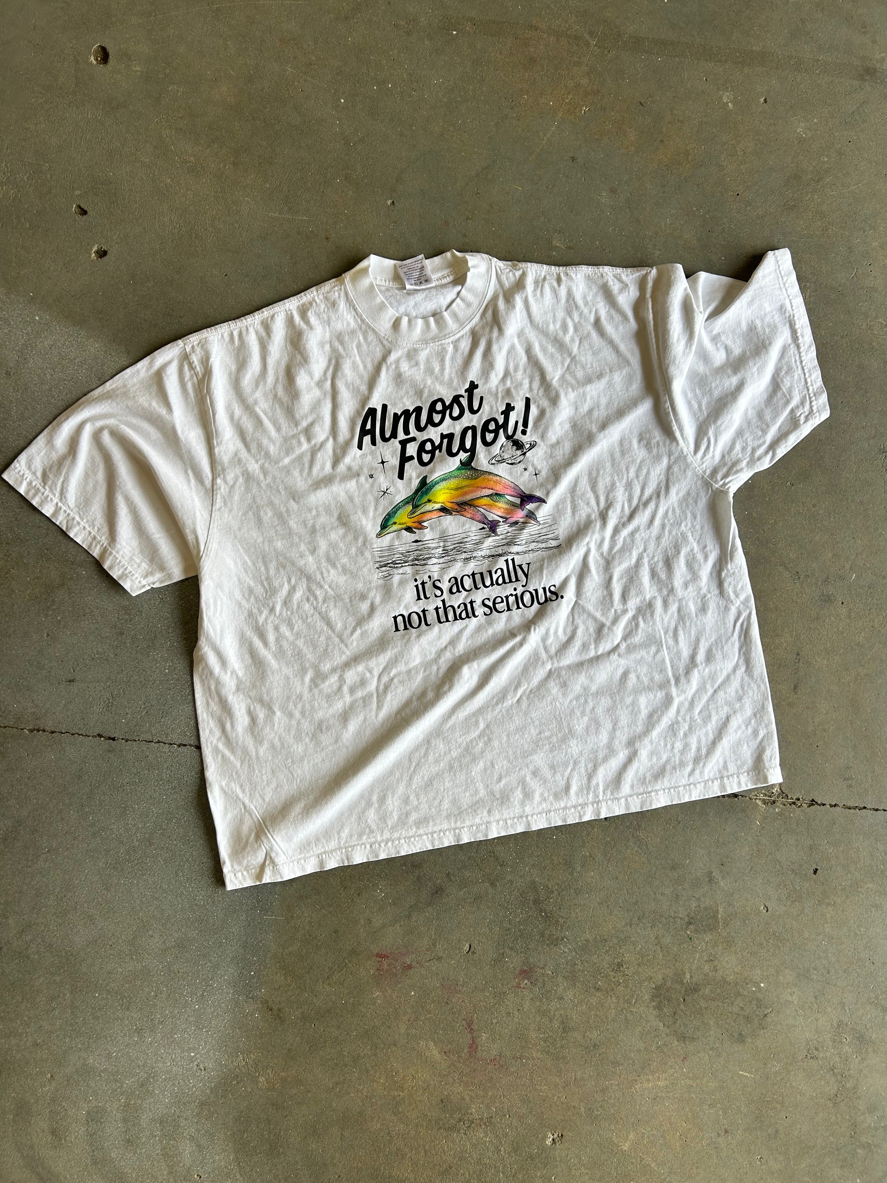 A white short-sleeve graphic t-shirt laid flat on a concrete floor. The shirt features a colorful fish illustration and the text 'Almost Forgot! It's actually not that serious' printed on the front.