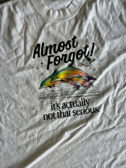 A close-up of a white graphic t-shirt featuring a colorful illustration of a dolphin jumping with the text 'Almost Forgot! it's actually not that serious.'.