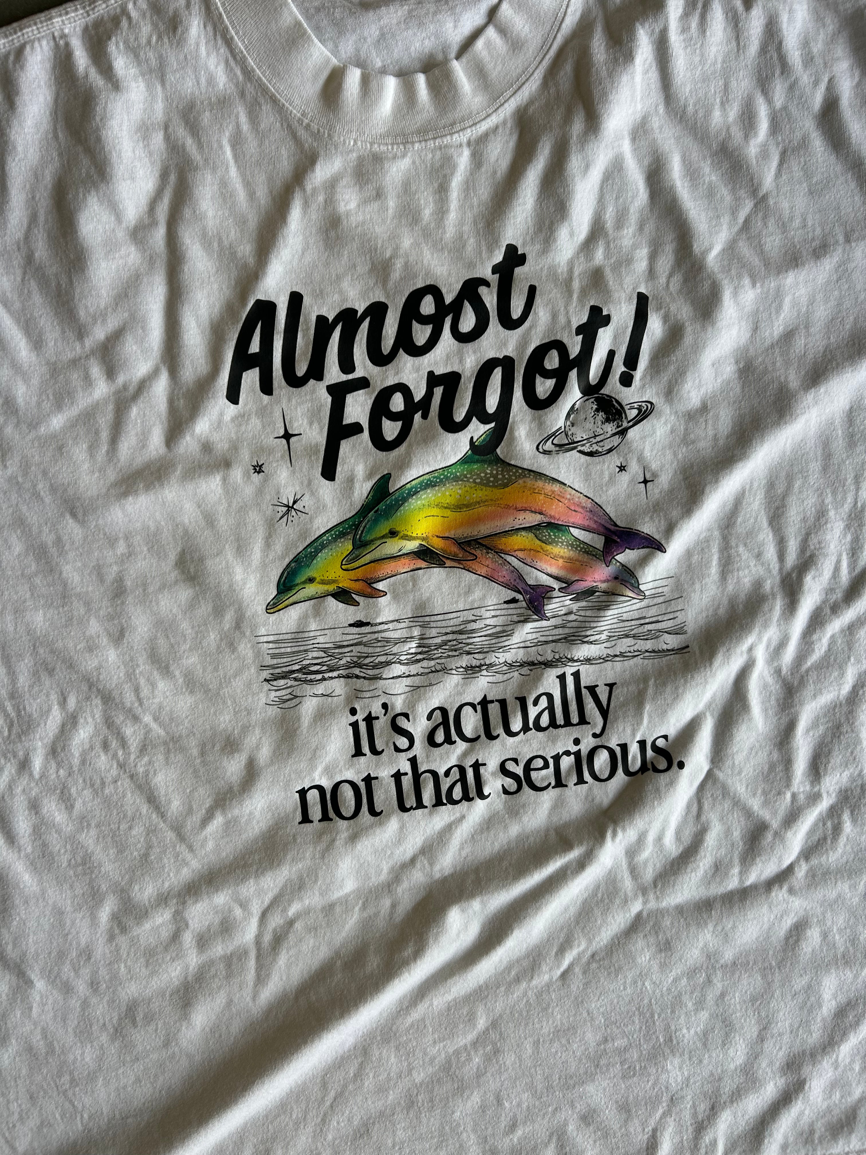 A close-up of a white graphic t-shirt featuring a colorful illustration of a dolphin jumping with the text 'Almost Forgot! it's actually not that serious.'.