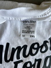Close-up of a white cotton garment tag reading 'SHAWK WEAR' and 'DROP SHOULDER GARMENT DYE' with size '2XL', attached to a white T-shirt with large black lettering.
