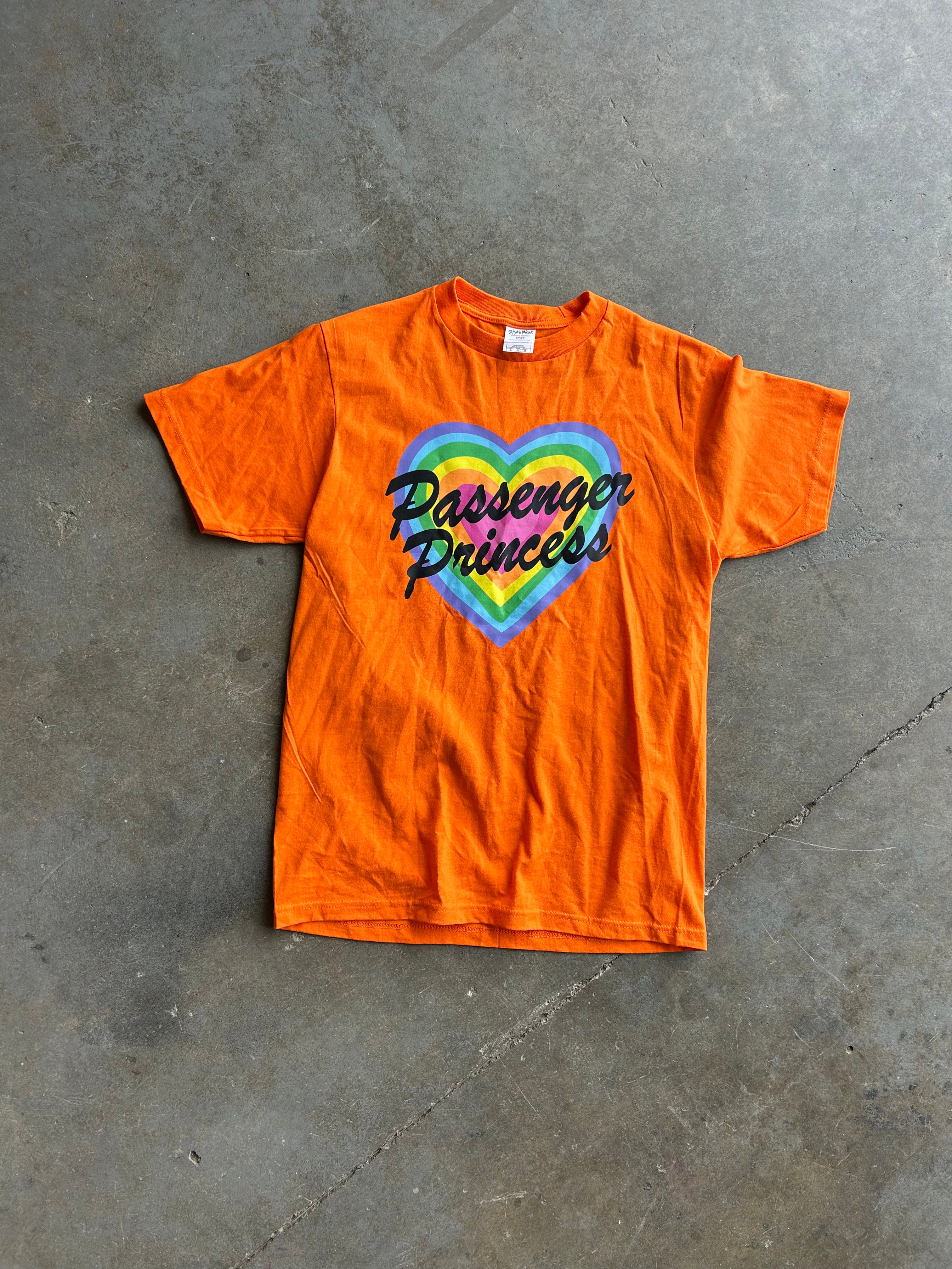 An orange short-sleeve t-shirt laid flat on a gray concrete floor, featuring a colorful rainbow heart graphic with the text 'Passenger Princess' in black script.