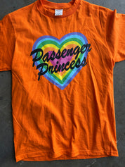 An orange short-sleeve t-shirt laid flat on a dark gray surface, featuring a graphic of a rainbow-striped heart with the words 'Passenger Princess' written in black script across the front.