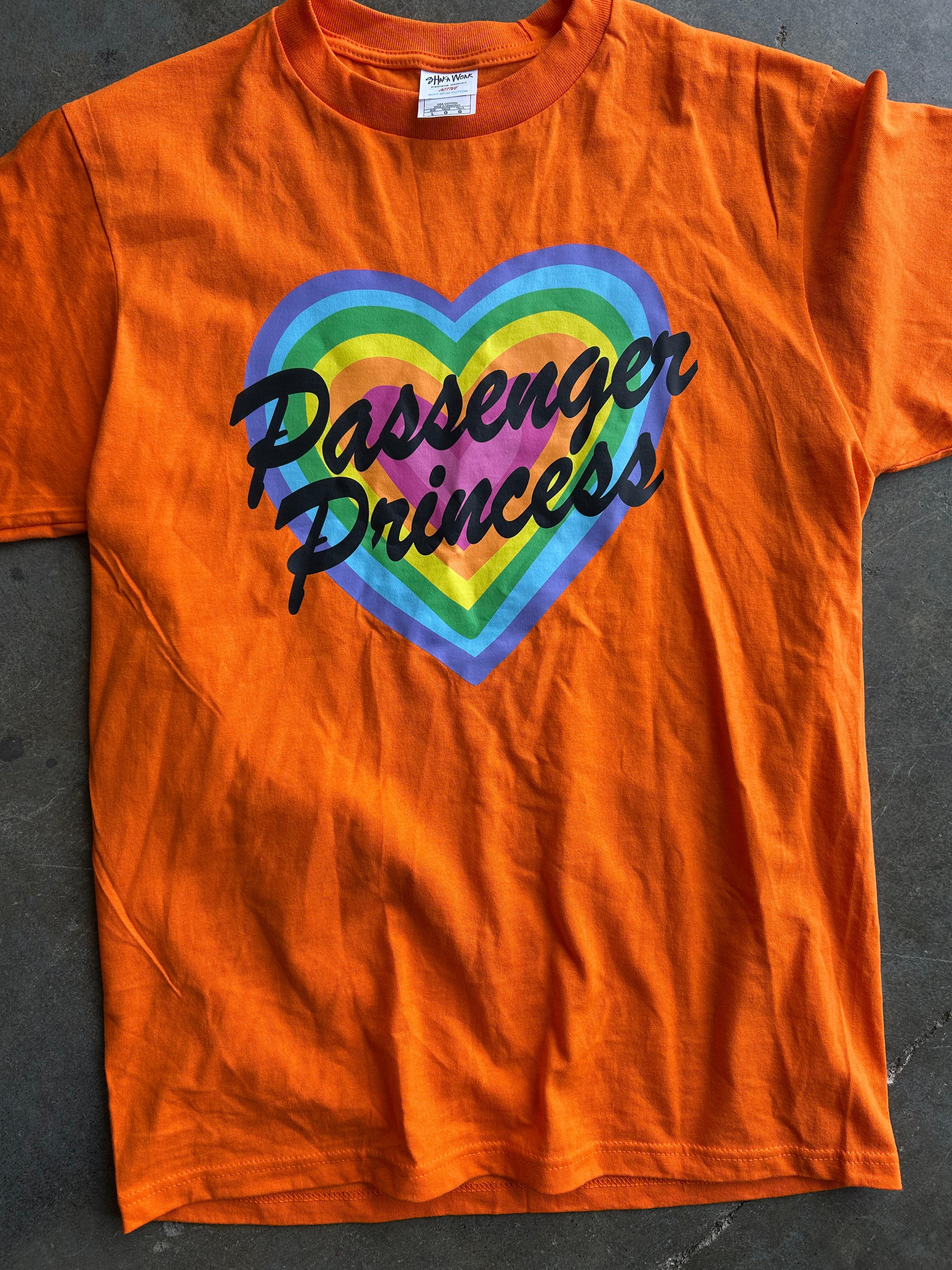 An orange short-sleeve t-shirt laid flat on a dark gray surface, featuring a graphic of a rainbow-striped heart with the words 'Passenger Princess' written in black script across the front.