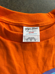 Close-up of the inside collar of an orange t-shirt showing the brand label 'Shaka Wear' and 'Active Soft Blend Cotton' on a dark gray textured background.
