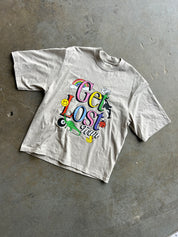 A light beige short-sleeve t-shirt with a colorful retro graphic reading 'Get Lost Fun' featuring cartoon elements, laid flat on a gray concrete floor.