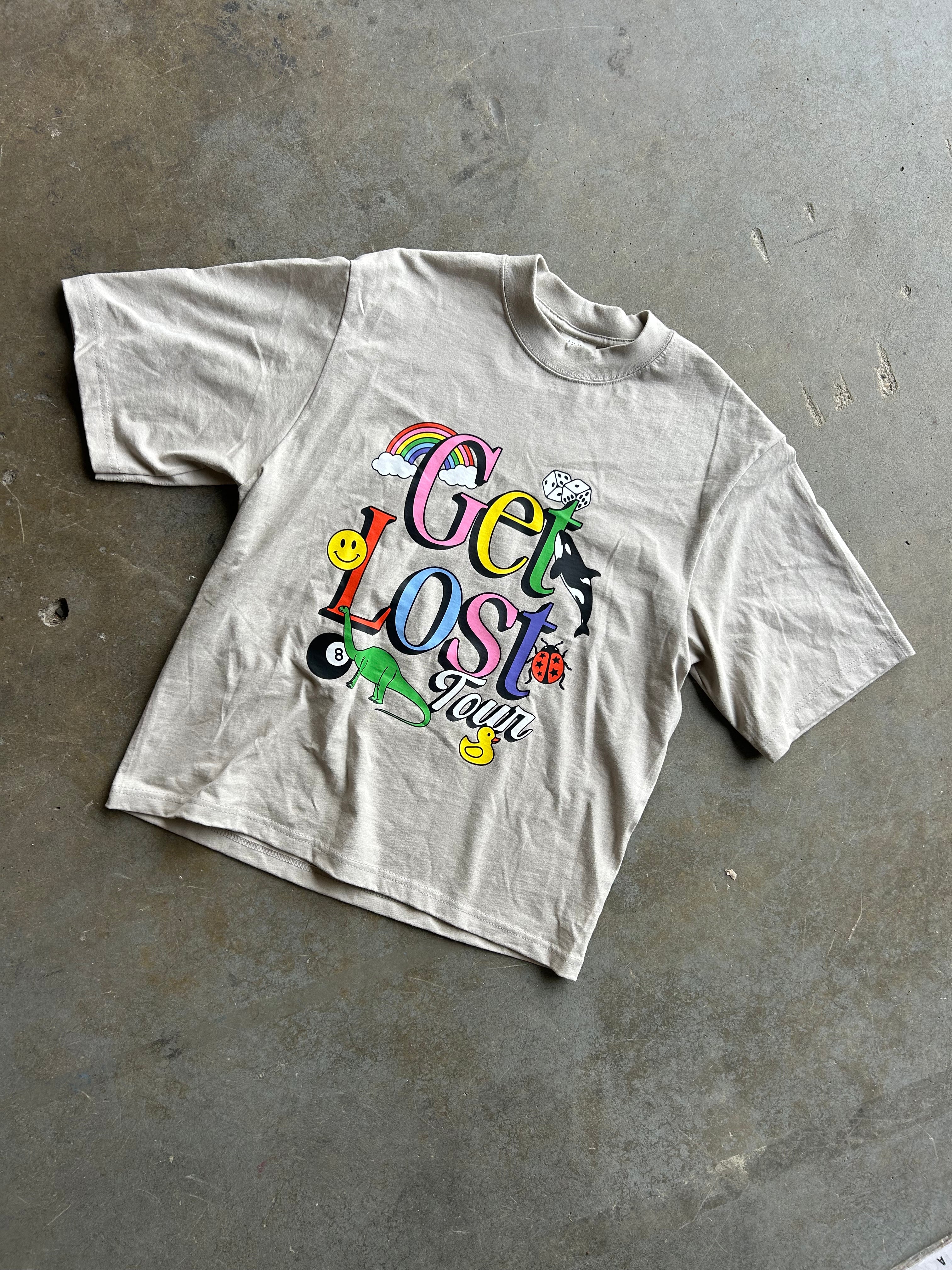 A light beige short-sleeve t-shirt with a colorful retro graphic reading 'Get Lost Fun' featuring cartoon elements, laid flat on a gray concrete floor.