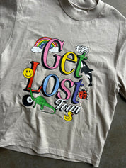 A light gray crewneck t-shirt featuring a colorful graphic print on the front with the words 'Get Lost Your' in stylized letters, accompanied by cartoon illustrations including a rainbow, smiley face, duck, ladybug, dinosaur, and orca.