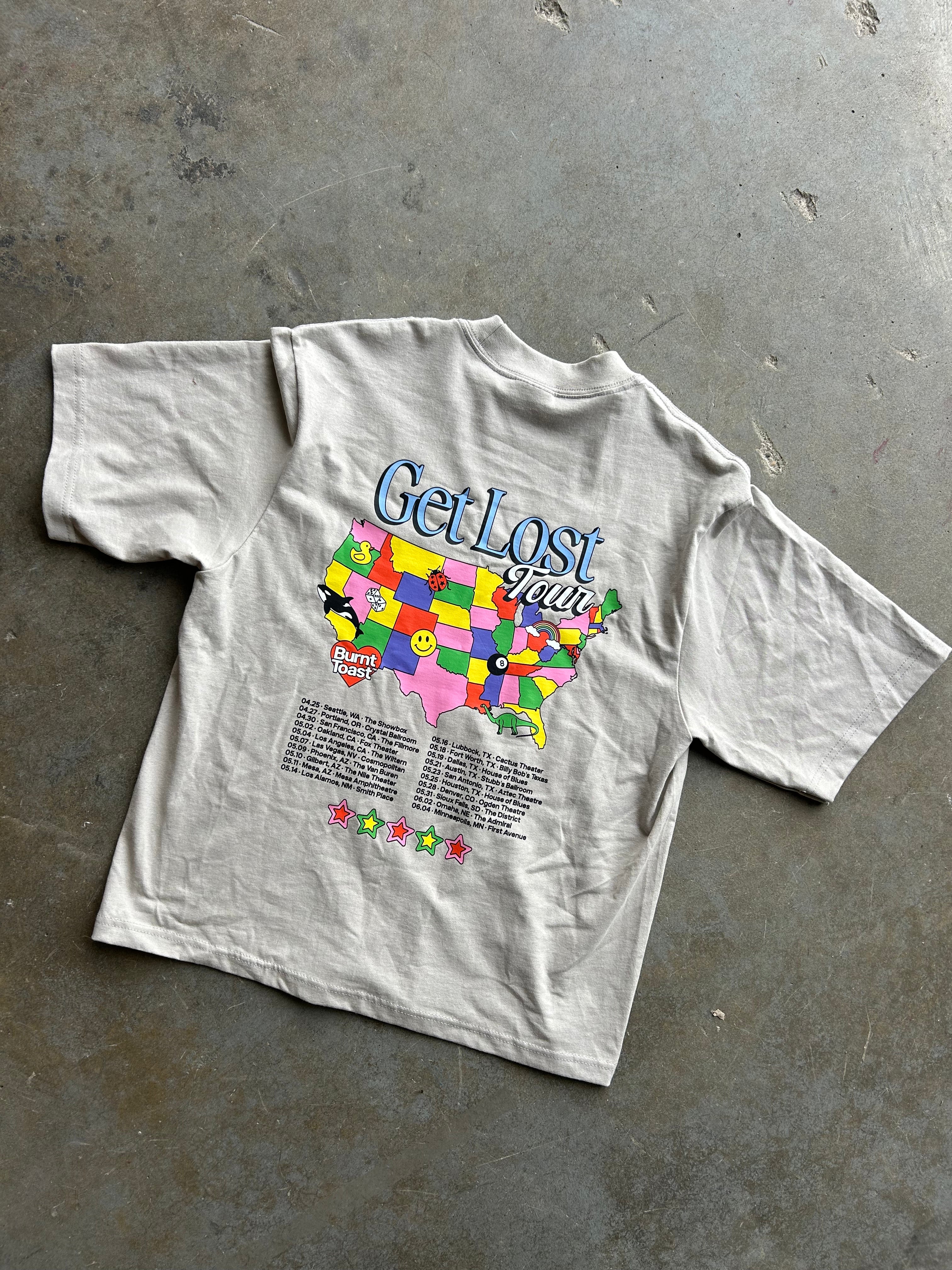 A light gray vintage t-shirt laid flat on a textured concrete floor, featuring a colorful graphic of the United States map with the text 'Get Lost Again' and a list of song titles below.