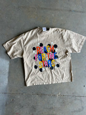 A beige short-sleeve graphic t-shirt laid flat on a gray concrete floor. The shirt features a colorful graphic on the front with the text 'REACH FOR THE STARS' in multicolored letters, surrounded by black stars and black dots.