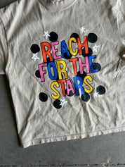 A beige cropped t-shirt featuring a colorful graphic that reads 'REACH FOR THE STARS' in multicolored letters, surrounded by black polka dots and white stars, laid flat on a gray concrete floor.