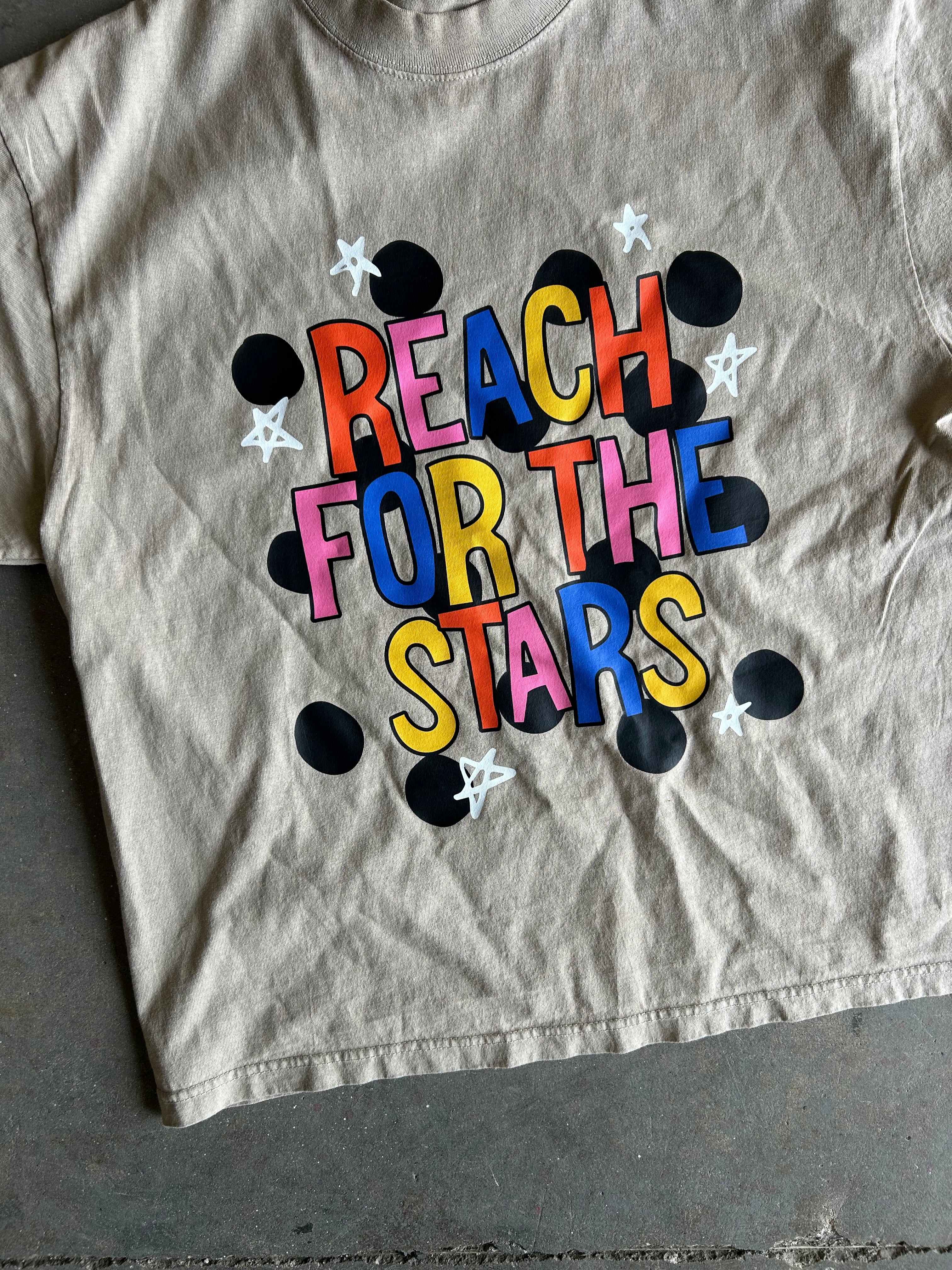 A beige cropped t-shirt featuring a colorful graphic that reads 'REACH FOR THE STARS' in multicolored letters, surrounded by black polka dots and white stars, laid flat on a gray concrete floor.